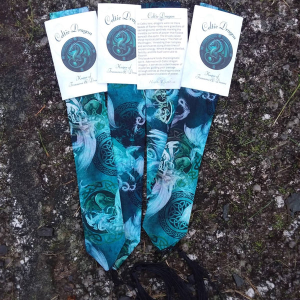 A pair of printed bookmarks with black tassels, laid flat on a textured dark ground. Each bookmark features a teal and blue fantasy-style illustration of dragons with Celtic knot patterns.