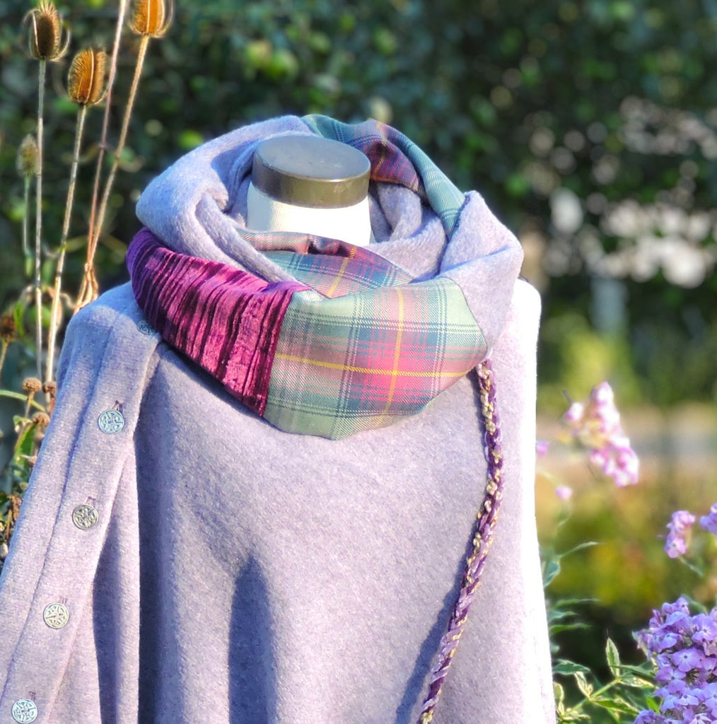 A light lavender wrap poncho draped on a mannequin outdoors, styled with a multicolor plaid scarf, a black crossbody bag with a plaid accent panel, and a silver-toned chain strap, set against a background of green foliage and purple flowers.