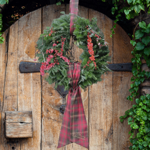 A green evergreen wreath with red berries and a Maple Leaf Tartan ribbon bow hanging on a rustic wooden door, with ivy visible around the edges.