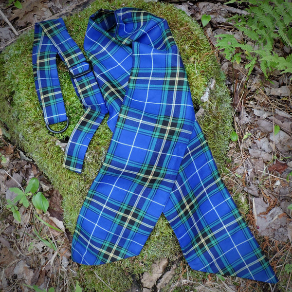 A NS tartan wreath sash and matching hanger laid out on a mossy log outdoors, surrounded by leaves and green plants.
